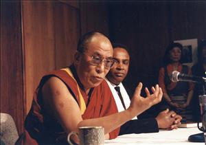 Seattle Mayor Norm Rice listens to Dalai Lama speak, with both sitting at a table on which there is a book, a coffee cup, and a microphone, with an Asian woman visible in background